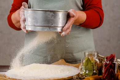 Person sifting flour into a bowl with a rustic kitchen setting.