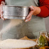 Person sifting flour into a bowl with a rustic kitchen setting.