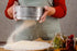 Person sifting flour into a bowl with a rustic kitchen setting.